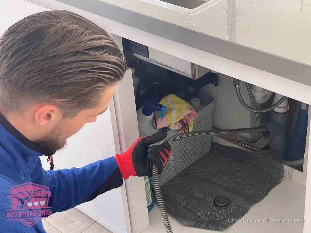 Een monteur voert een flexibele ontstoppingsveer in onder het keukenkastje, langs de sifon, om de gootsteen te ontstoppen in Heerenveen