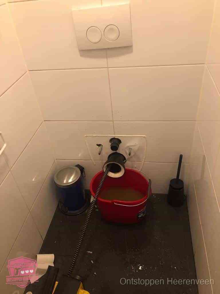 Een ontstopte wand-wc met verwijderde toiletpot waar een ontstoppingsveer in de wanduitgang verdwijnt, het achterhalen waar de verstopping zit in Heerenveen