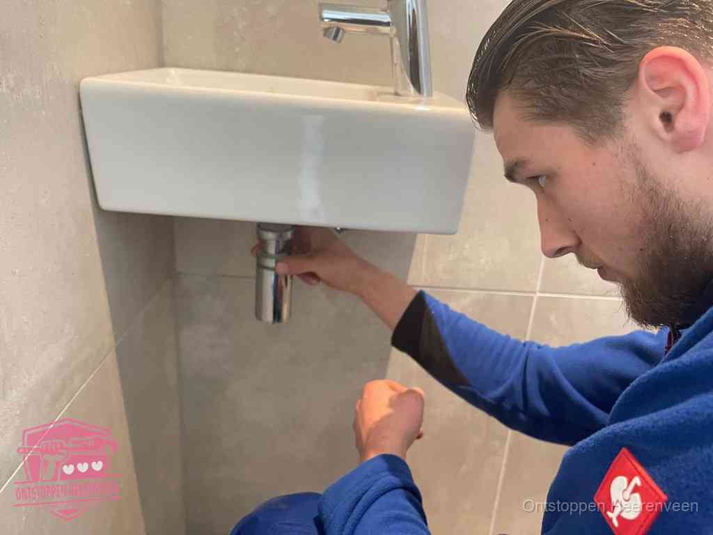 Een monteur draait met de hand de zwanenhalssifon onder een fonteinwasbak in de toiletruimte los om de bron van rioollucht in Heerenveen te vinden